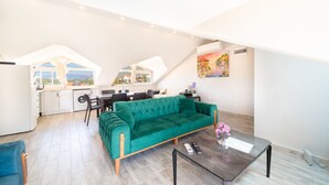 En moderne loftlejlighed med en grøn sofa, spiseområde, køkken og et stort vindue med udsigt over landskabet.
