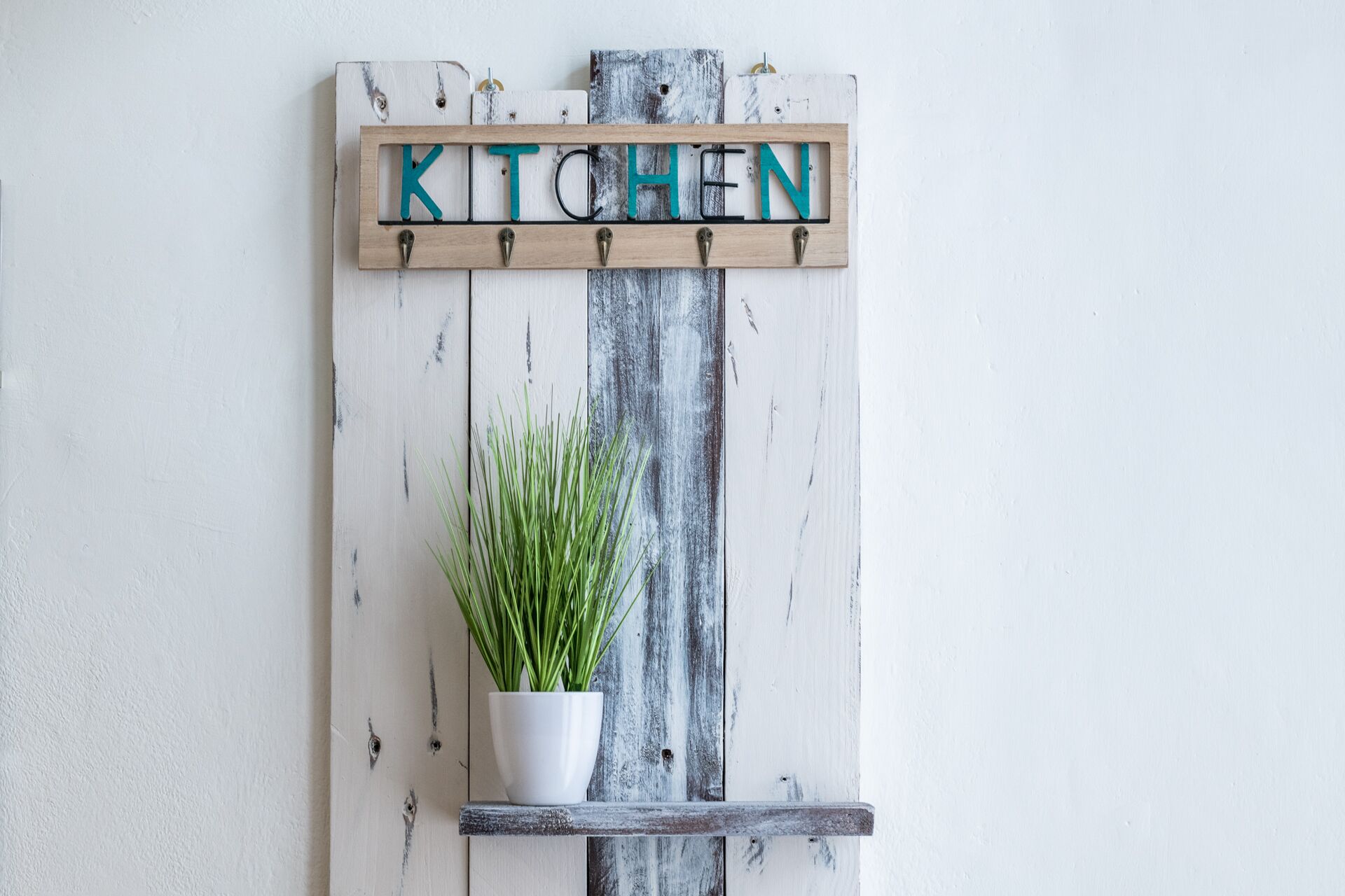 Un letrero de madera rústica con la palabra "KITCHEN" en letras azul verdosas, sujeto a una pared, con una planta en maceta debajo.