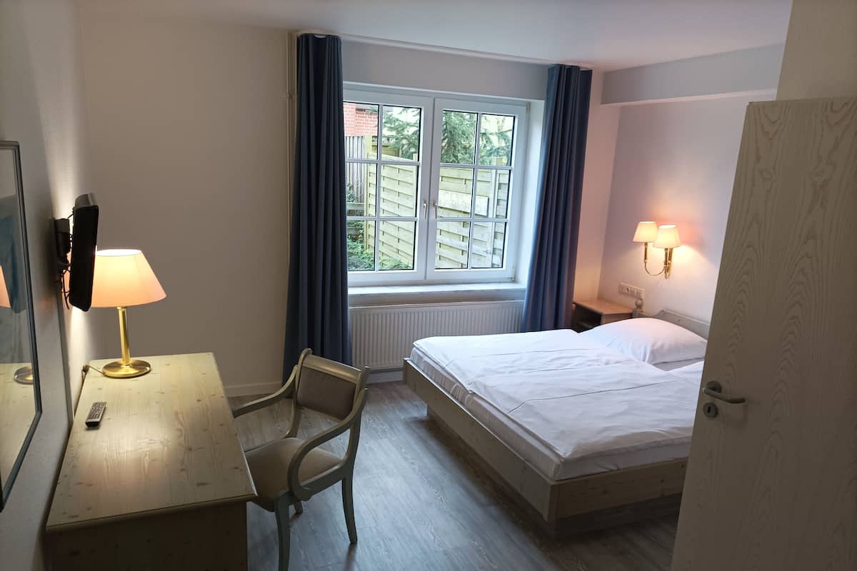 Una habitación de hotel con una cama, un escritorio con lámpara, una silla, una ventana con cortinas y una mesita de noche con lámpara.