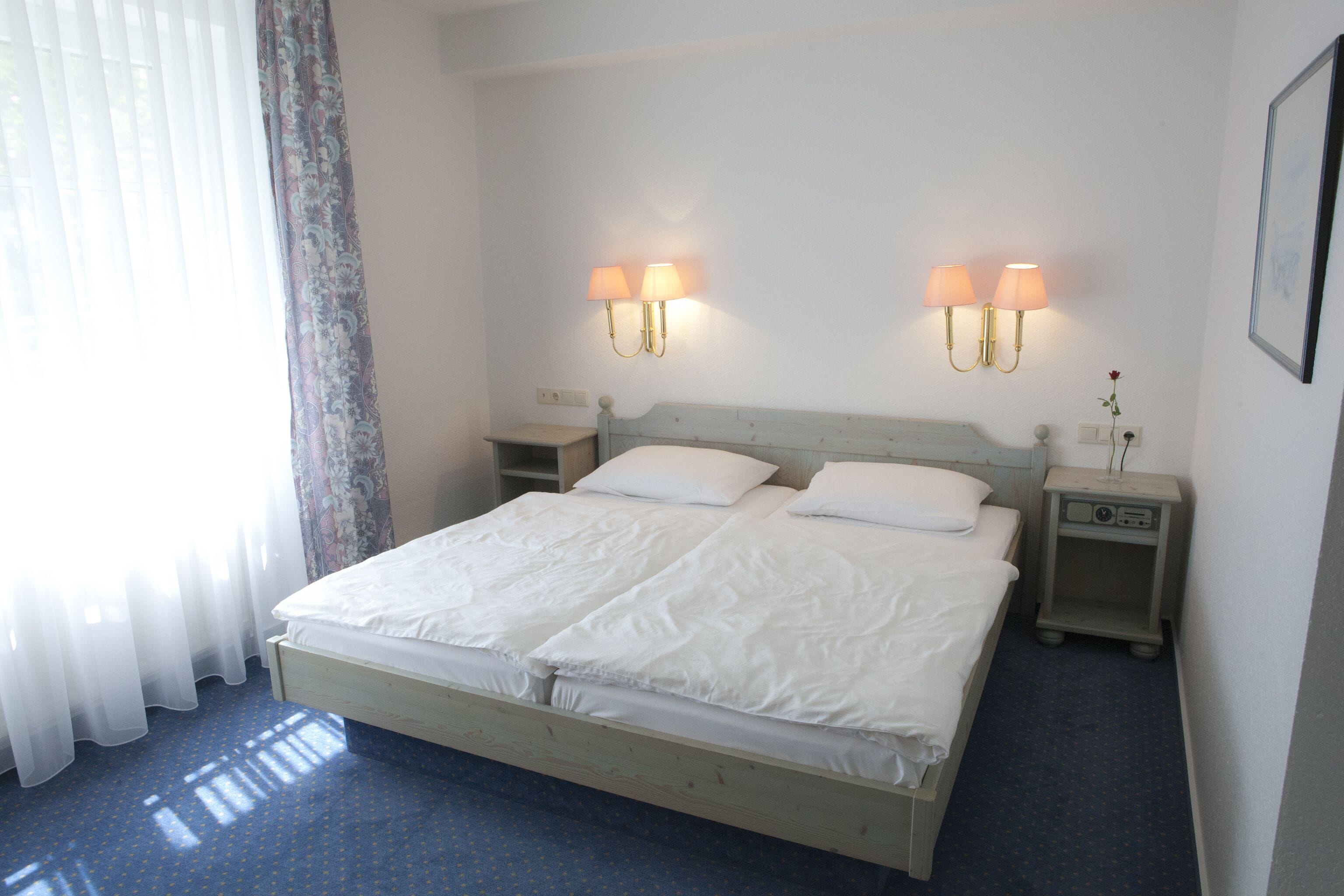 A double bed with white linens, two bedside tables with lamps, a nightstand with a clock, and a window with sheer curtains.