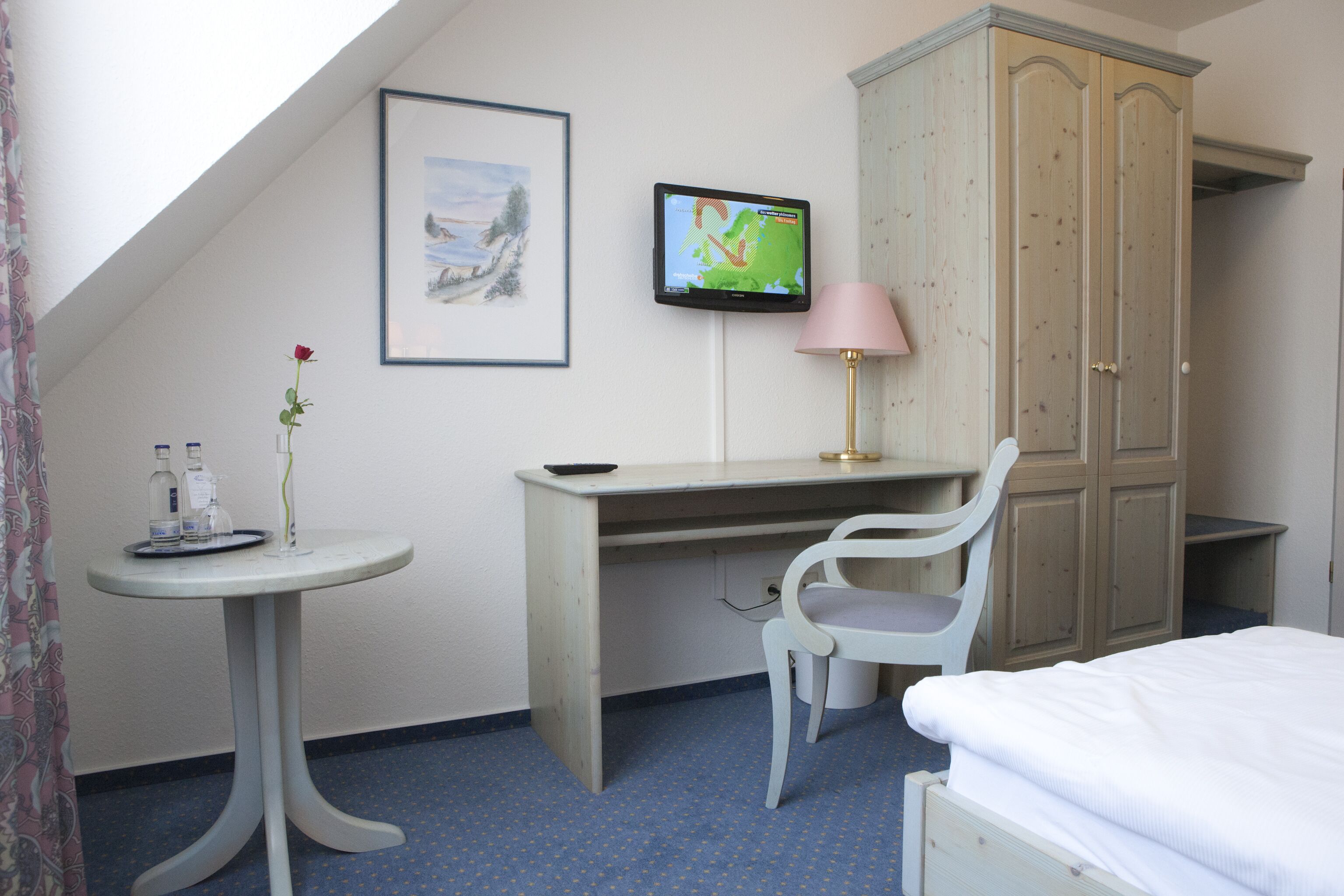 Habitación de hotel con cama, escritorio, silla, televisor, armario y una pequeña mesa con un jarrón de flores.