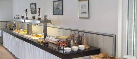 A buffet setup with various food items and beverages, a cutting board with utensils, and framed artwork on the wall.