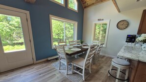Un comedor con mesa y sillas de madera, ventanas amplias y una zona de cocina con un reloj y una cafetera.