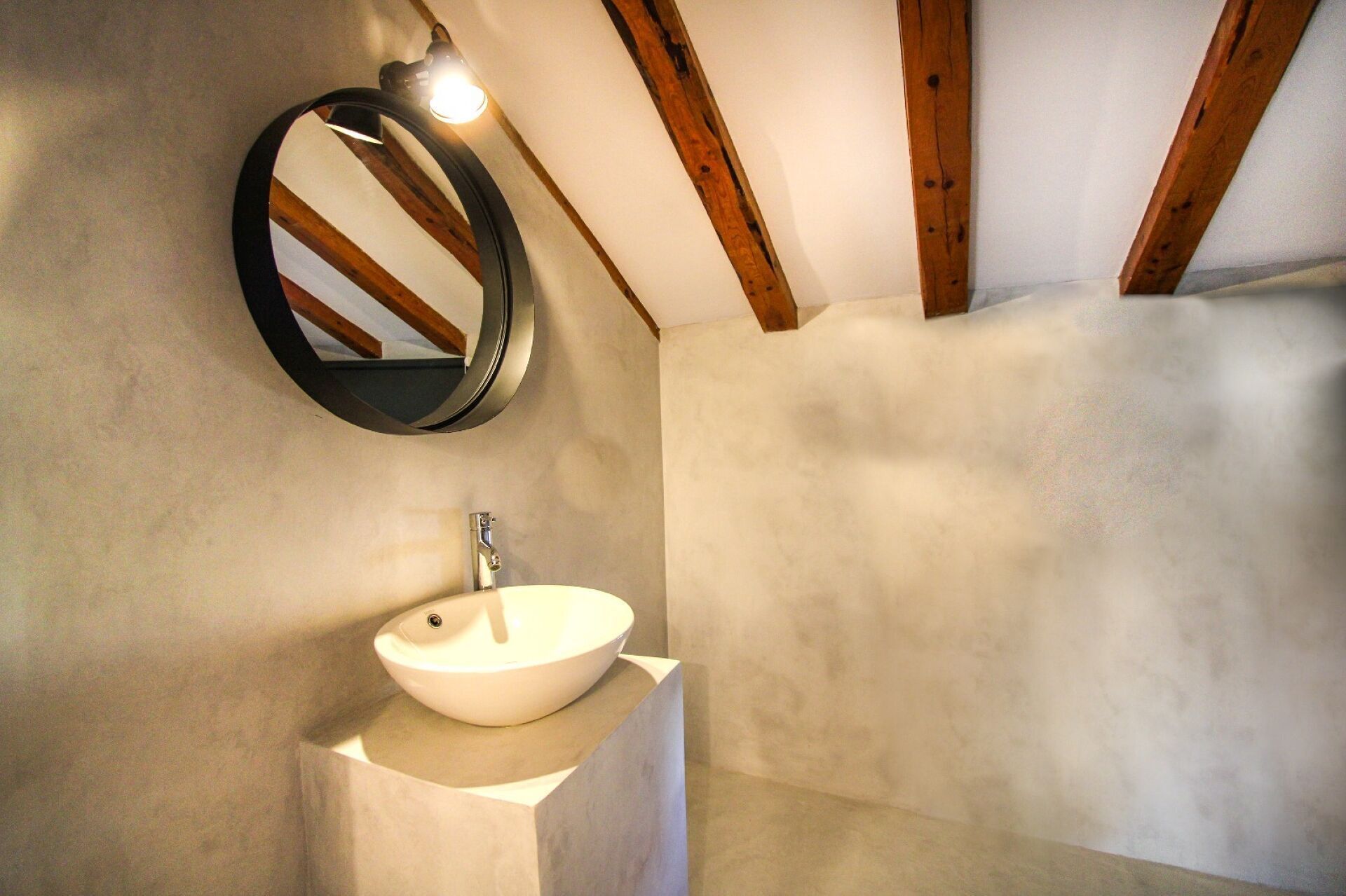 Une salle de bain avec un miroir rond, un lavabo blanc et des poutres en bois au plafond.