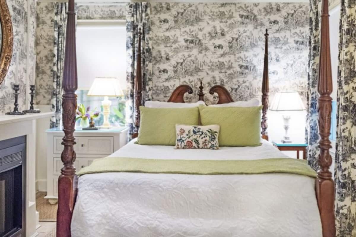 A four-poster bed with green and white bedding, a floral-patterned headboard, and a nightstand with a lamp.