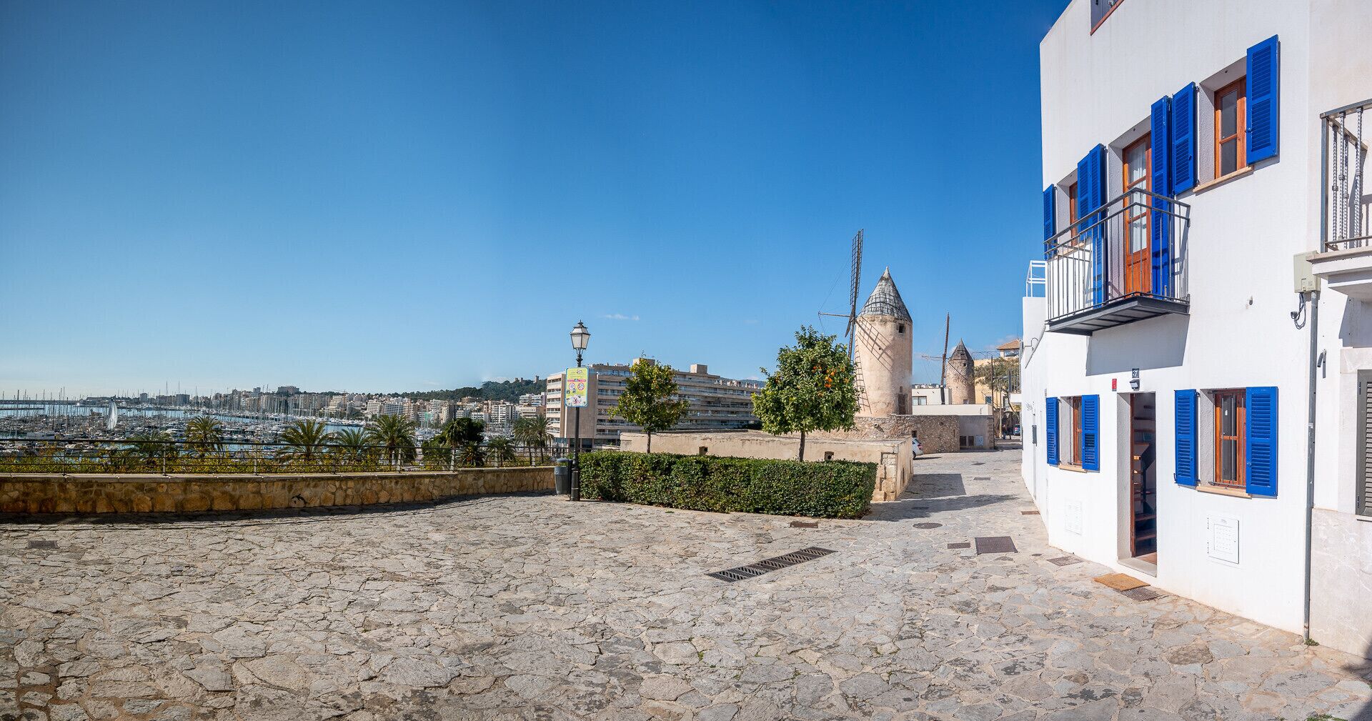 Ein mit Pflastersteinen belegter Innenhof mit Blick auf einen Yachthafen und eine Stadtlandschaft, einen Turm sowie Gebäude mit blauen Fensterläden.