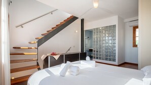 Habitación de hotel con cama, escalera de madera, baño con ducha de vidrio y vistas al exterior.