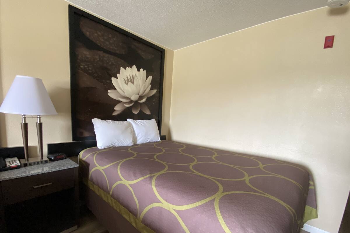 A hotel room with a bed, a desk with a lamp, and a large floral artwork on the wall.