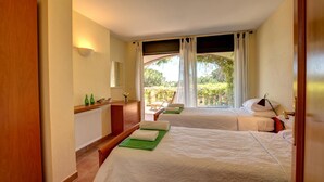 A bedroom with two beds, a wooden wardrobe, a bedside table with a mirror, and a view of greenery outside.