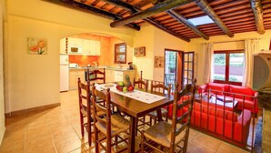 A dining area with a wooden table and chairs, a kitchen in the background, and a red sofa in the foreground. - Níspero - Two Bedroom Villa, Sleeps 4 (Begur)