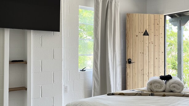 A bedroom with a bed, a wooden wardrobe, a flat-screen TV mounted on a white brick wall, and a window with curtains. - Triangle Motel (Red Bluff)