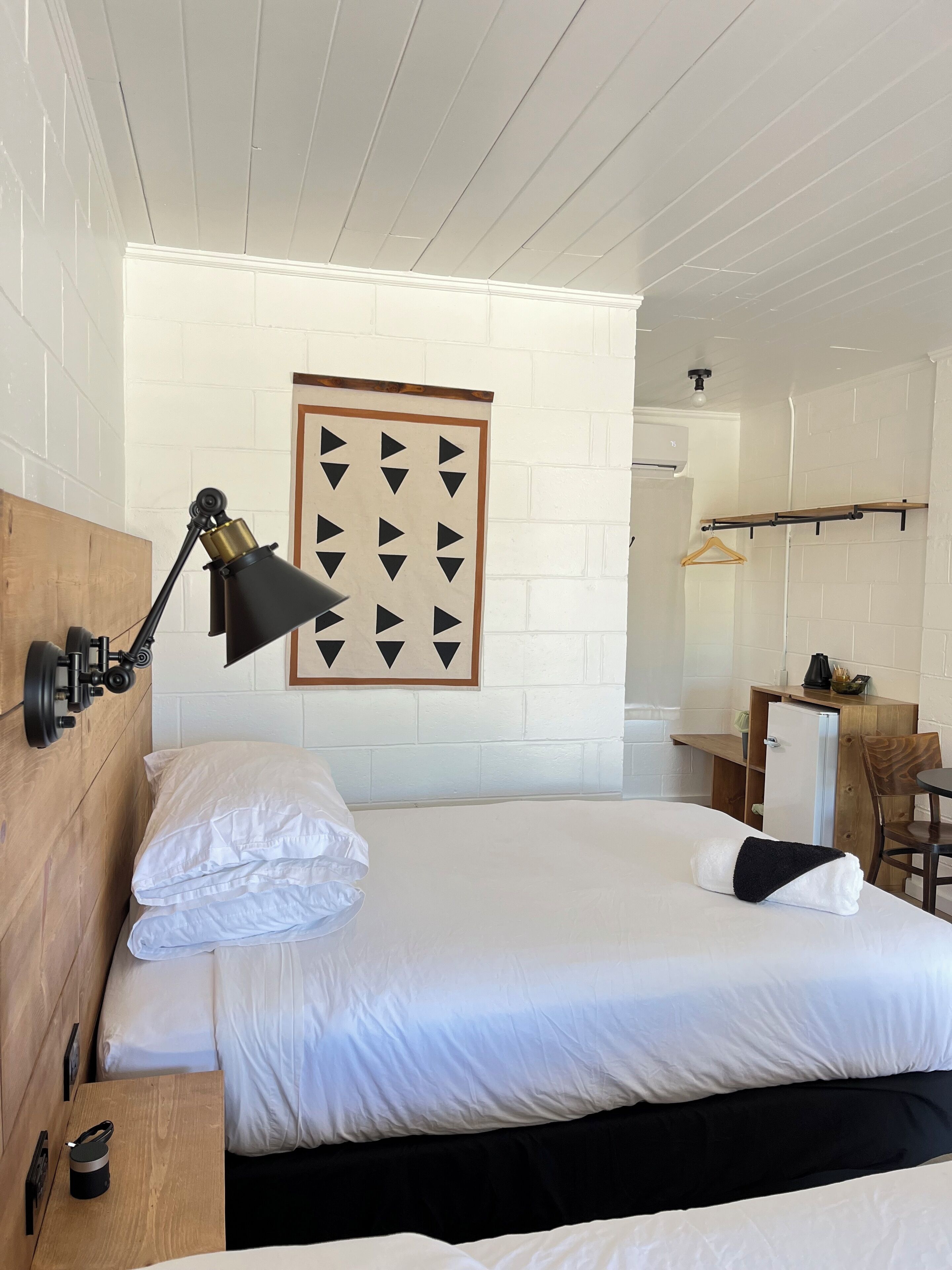 A hotel room with a bed, a desk, a chair, a hanging coat rack, and a wall-mounted lamp.