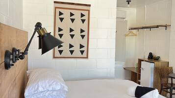 A hotel room with a bed, a desk, a chair, a hanging coat rack, and a wall-mounted lamp.