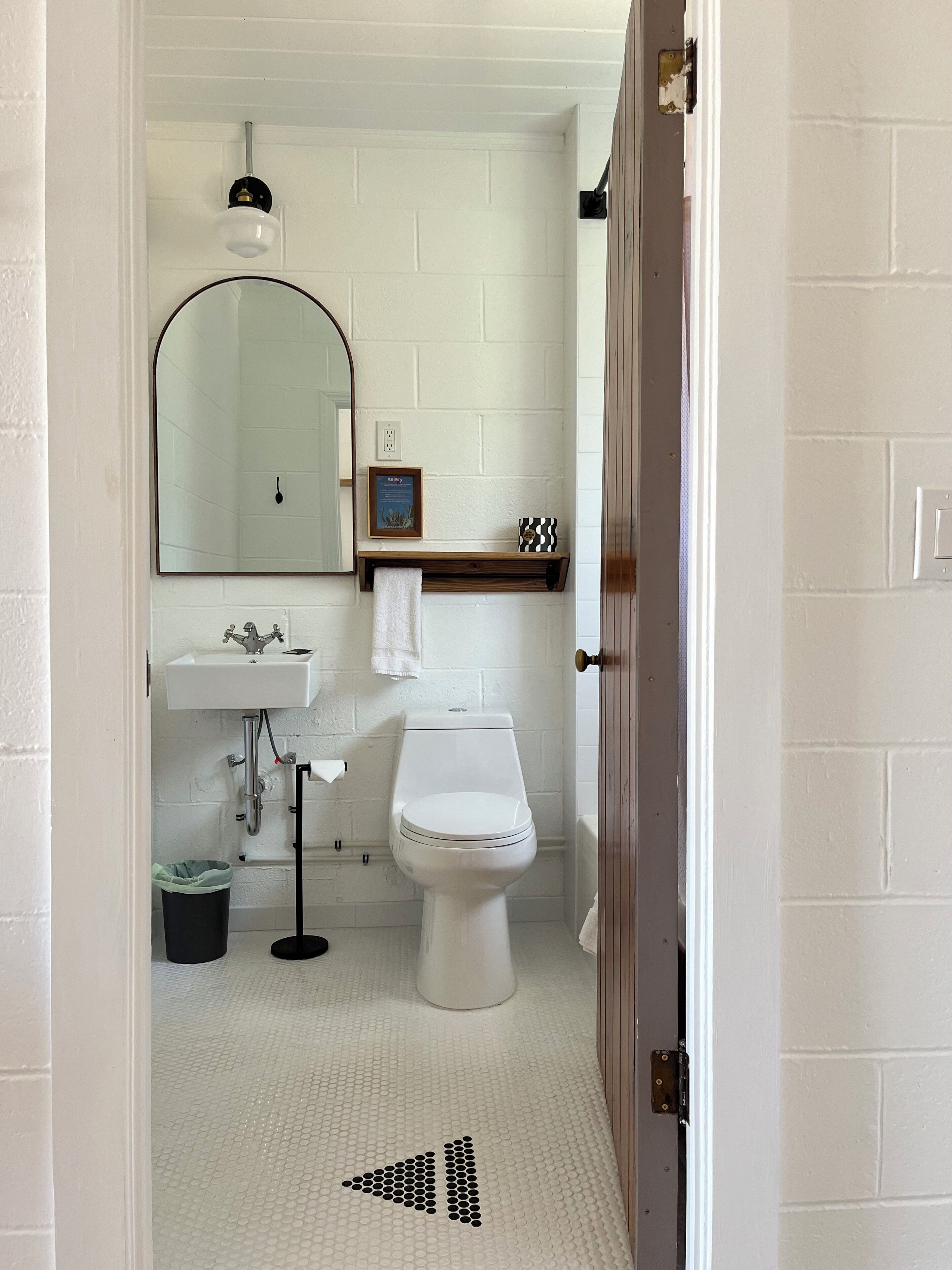 Une salle de bain avec des toilettes, un lavabo et un miroir, tous de couleur blanche. On y trouve une petite étagère avec un tableau encadré et une serviette. Le sol est décoré d'un motif à bulles.