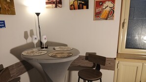 Un pequeño comedor con una mesa redonda, dos sillas y un taburete. Hay dos copas de vino sobre la mesa, y un cartel en la pared.