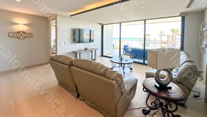 Un salon moderne avec un canapé beige, une table basse et un téléviseur. On peut voir une pancarte sur laquelle est écrit « MAISON DE PLAGE », et de grandes fenêtres offrent une vue sur la plage.