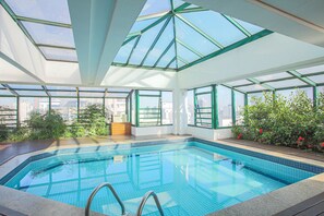 A swimming pool with a glass ceiling, surrounded by greenery and city views.