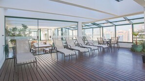 A sunlit room with a wooden floor, large windows, and several white cushioned chairs. - Edifício Expert (São Paulo)