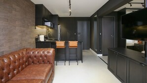 A modern kitchen with dark cabinetry, a leather sofa, and a flat-screen TV. - Edifício Expert (São Paulo)
