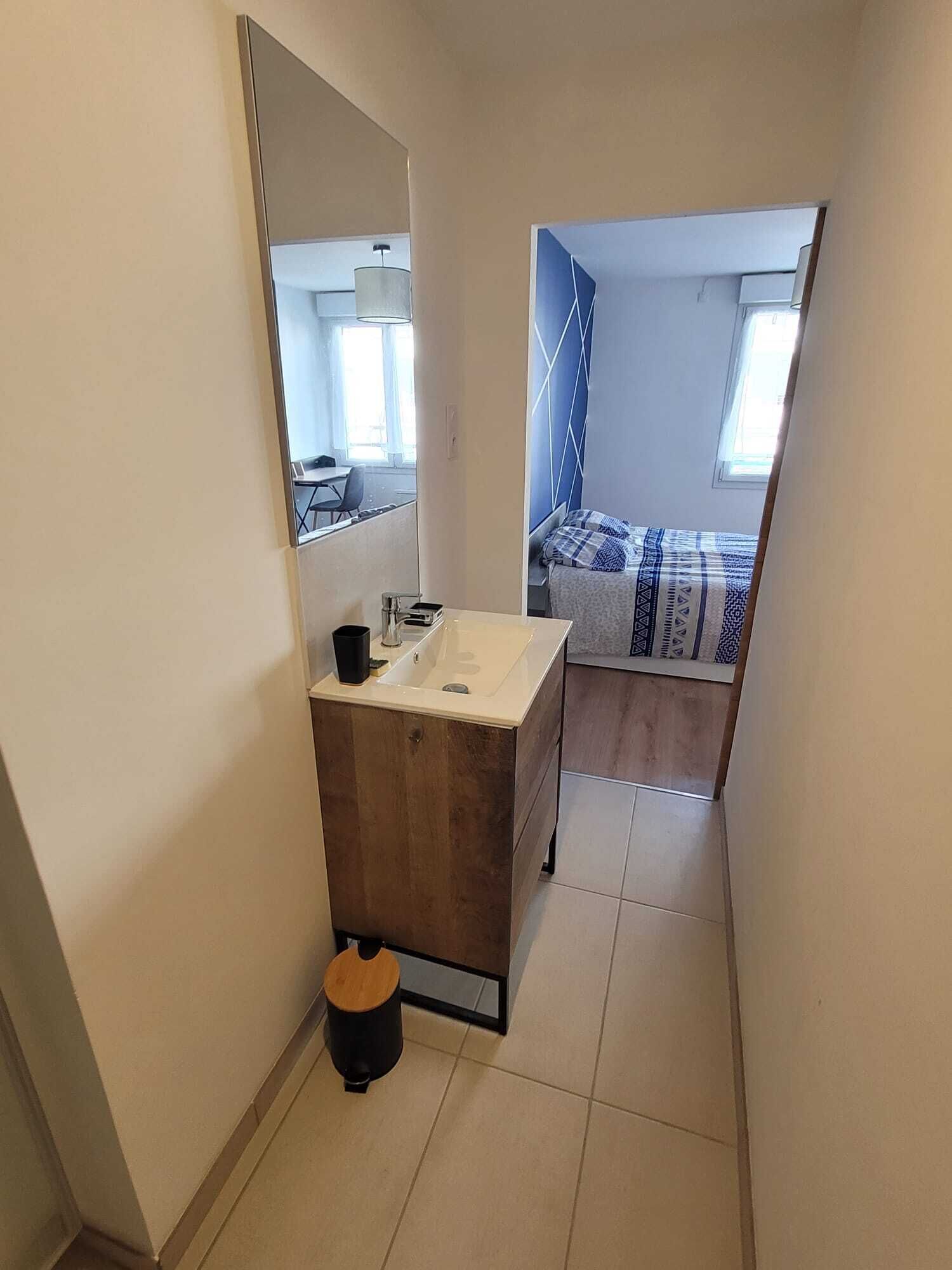 Um banheiro estreito com toucador de madeira, um espelho e um pequeno banquinho. Uma cama com colcha estampada é visível no quarto adjacente.