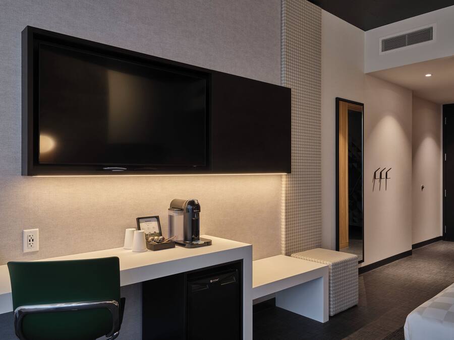 Quarto de hotel moderno com uma TV de tela plana grande na parede, uma pequena escrivaninha com cafeteira e uma cadeira verde.