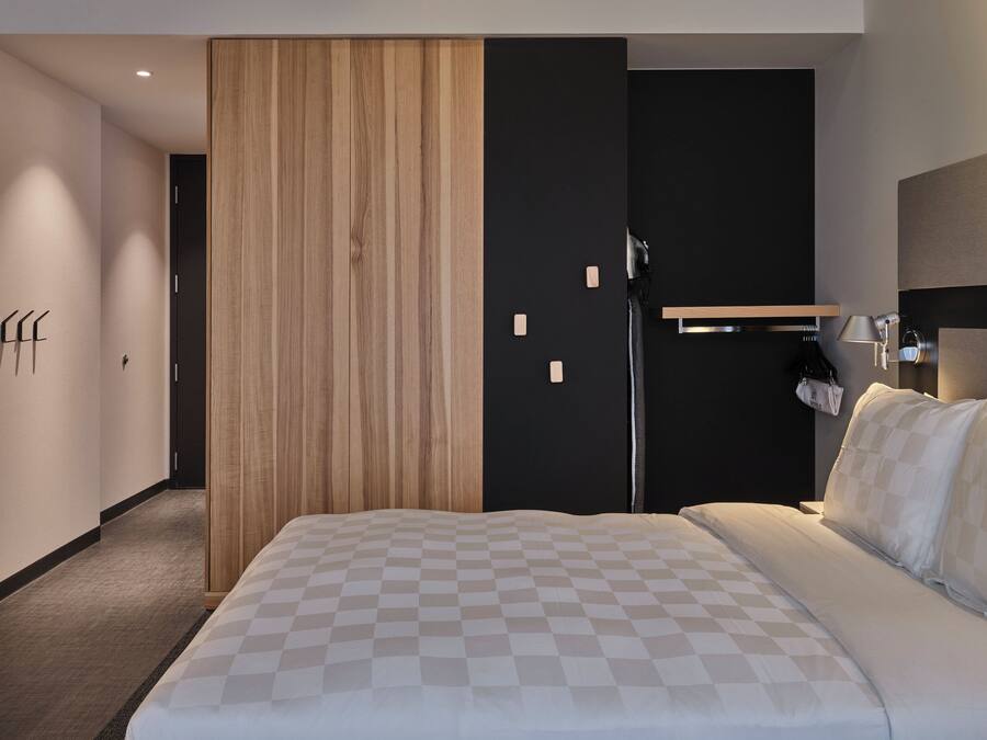 A modern hotel room with a large bed, a wooden wardrobe, a black door, and a small table with a lamp.
