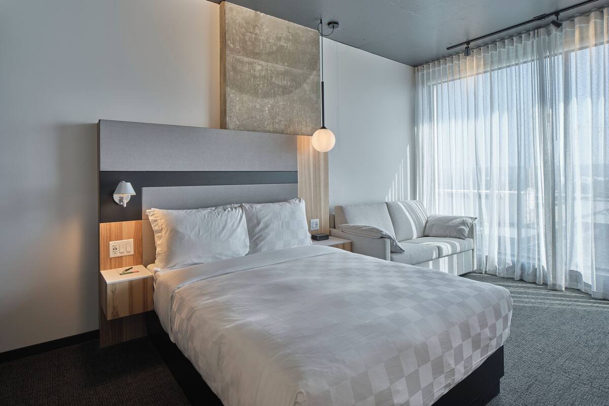 A modern hotel room with a large bed, a bedside table with a lamp, a small desk, and a chair.