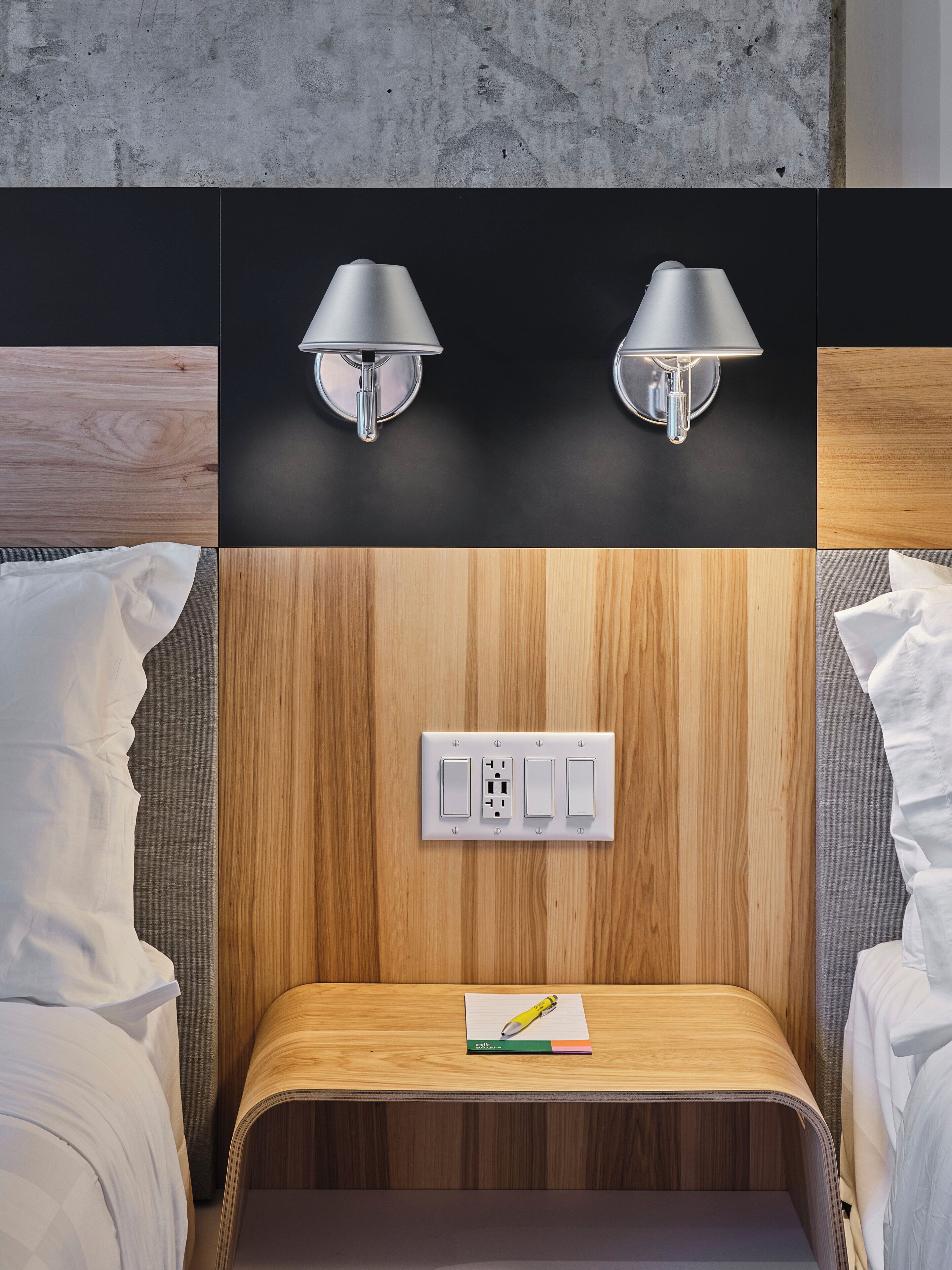 A hotel room with a wooden panel wall, two wall-mounted lamps, a power outlet, and a small wooden table with a notebook and pen.
