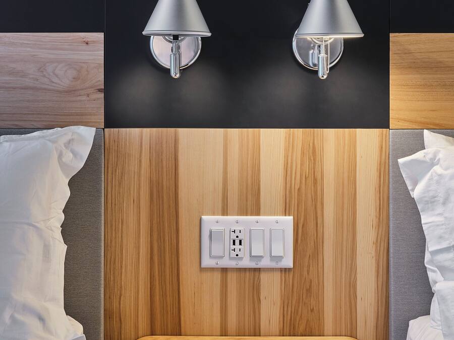 Hotelkamer met een wandpaneel van hout, twee wandlampen, een stopcontact en een klein houten tafeltje met een notitieboekje en pen.