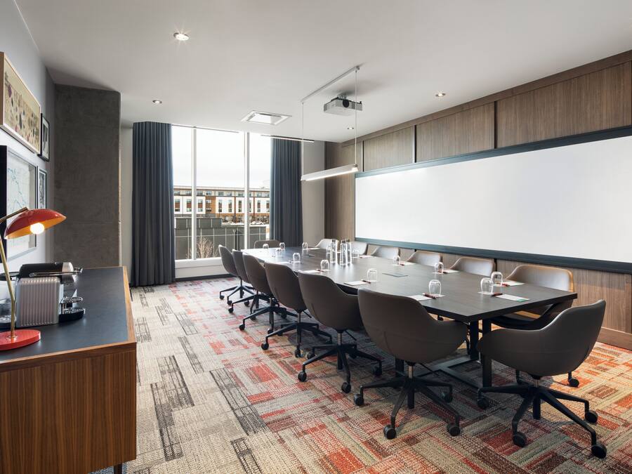 A conference room with a large table, chairs, a projector screen, and a view of buildings through large windows.