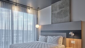 Une chambre d’hôtel moderne avec un grand lit, une table de chevet, une lampe et une vue sur la ville à travers des rideaux transparents.