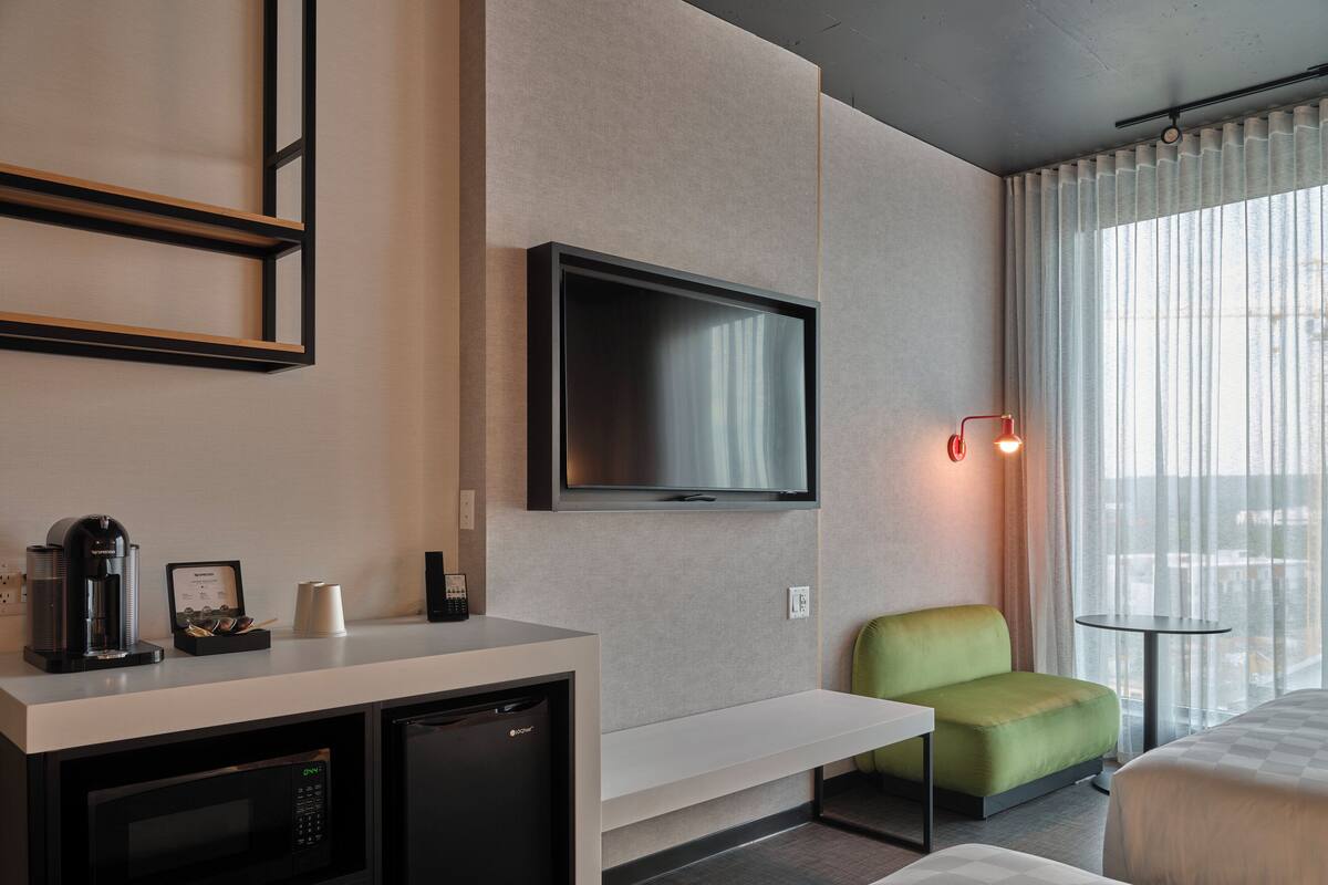 A modern hotel room with a kitchenette, a flat-screen TV mounted on the wall, a green armchair, and a bed with a checkered duvet.