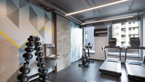 Une salle de sport équipée de matériel d’exercice, avec un distributeur d’eau et une fenêtre donnant sur des immeubles.