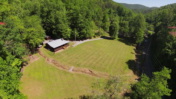 An aerial view of a property with a large lawn, a wooden cabin, and dense forested hills in the background.