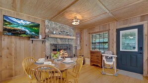 Un intérieur en bois avec une table à manger pour quatre personnes, une cheminée en pierre et un téléviseur fixé au mur.