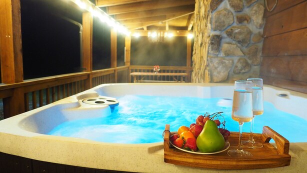Un bain à remous avec une terrasse en bois, un mur en pierre et un plateau contenant des fruits et des verres.