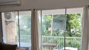 Ein Zimmer mit einem braunen Sofa, einem gemusterten Zierkissen und einem großen Fenster mit Blick auf einen Balkon und Grün.