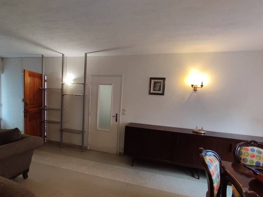 Habitació amb un sofà, una porta de fusta, un mirall amb marc blanc, un llum penjada a la paret i una taula de dinar amb cadires.