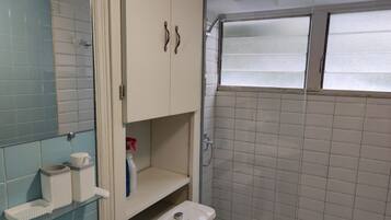 A bathroom with a toilet, a glass shelf with toiletries, a cabinet, and a shower area with frosted glass doors.