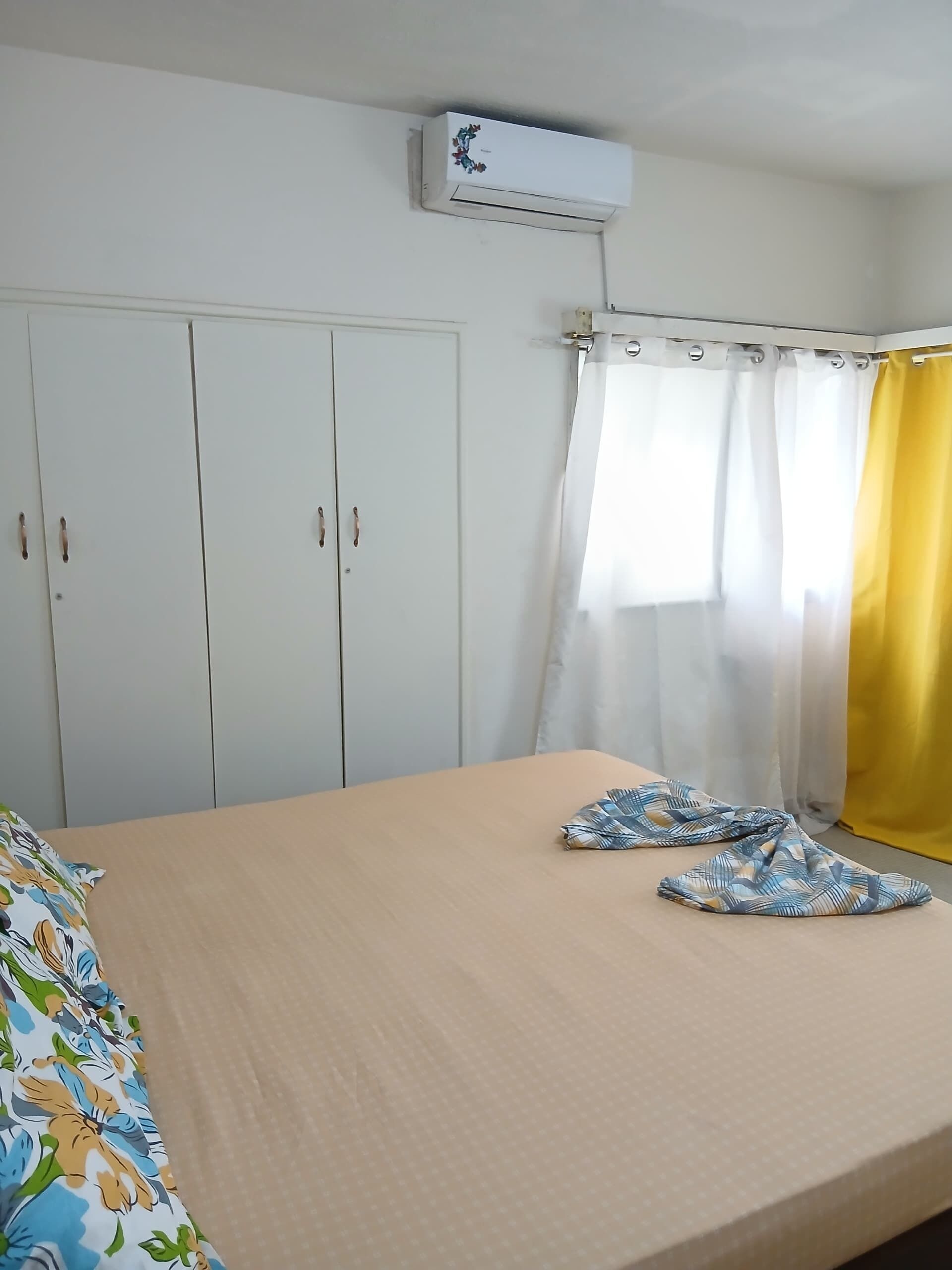A bedroom with a bed, a wardrobe, an air conditioning unit, and a window with curtains.