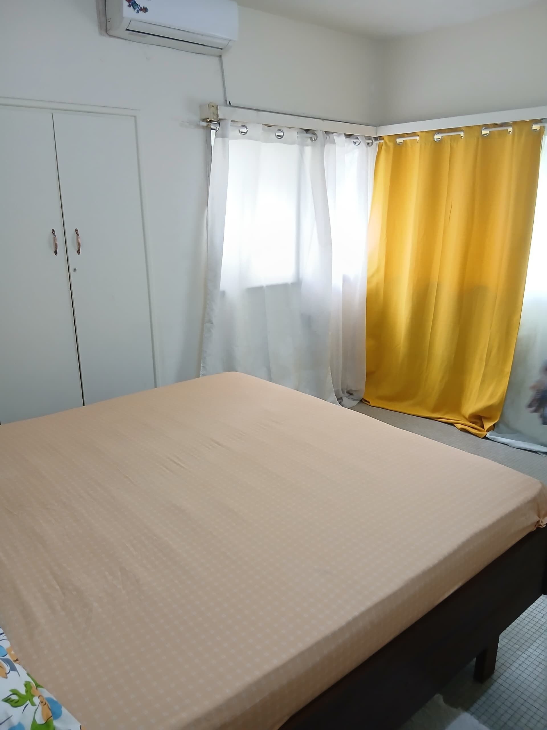 A room with a bed, a wardrobe, a window with curtains, and an air conditioning unit.