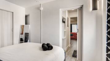 Habitación de hotel con una cama grande, una mesita de noche, un espejo y un armario. Se ve un pasillo que conduce a otra habitación.