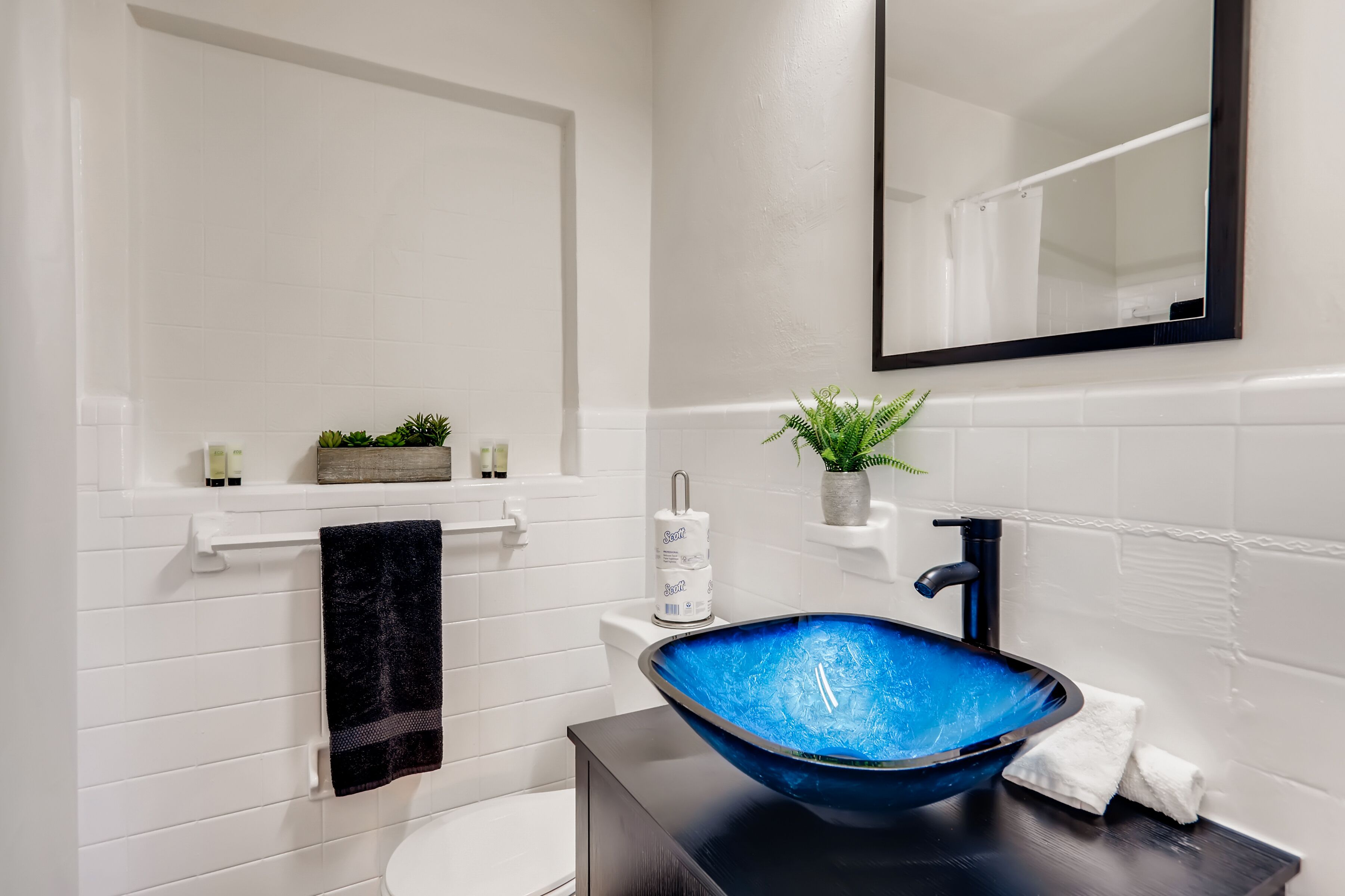 A bathroom with a white tiled wall, a black sink with a blue basin, a white toilet, a black towel, and a small plant on a shelf.