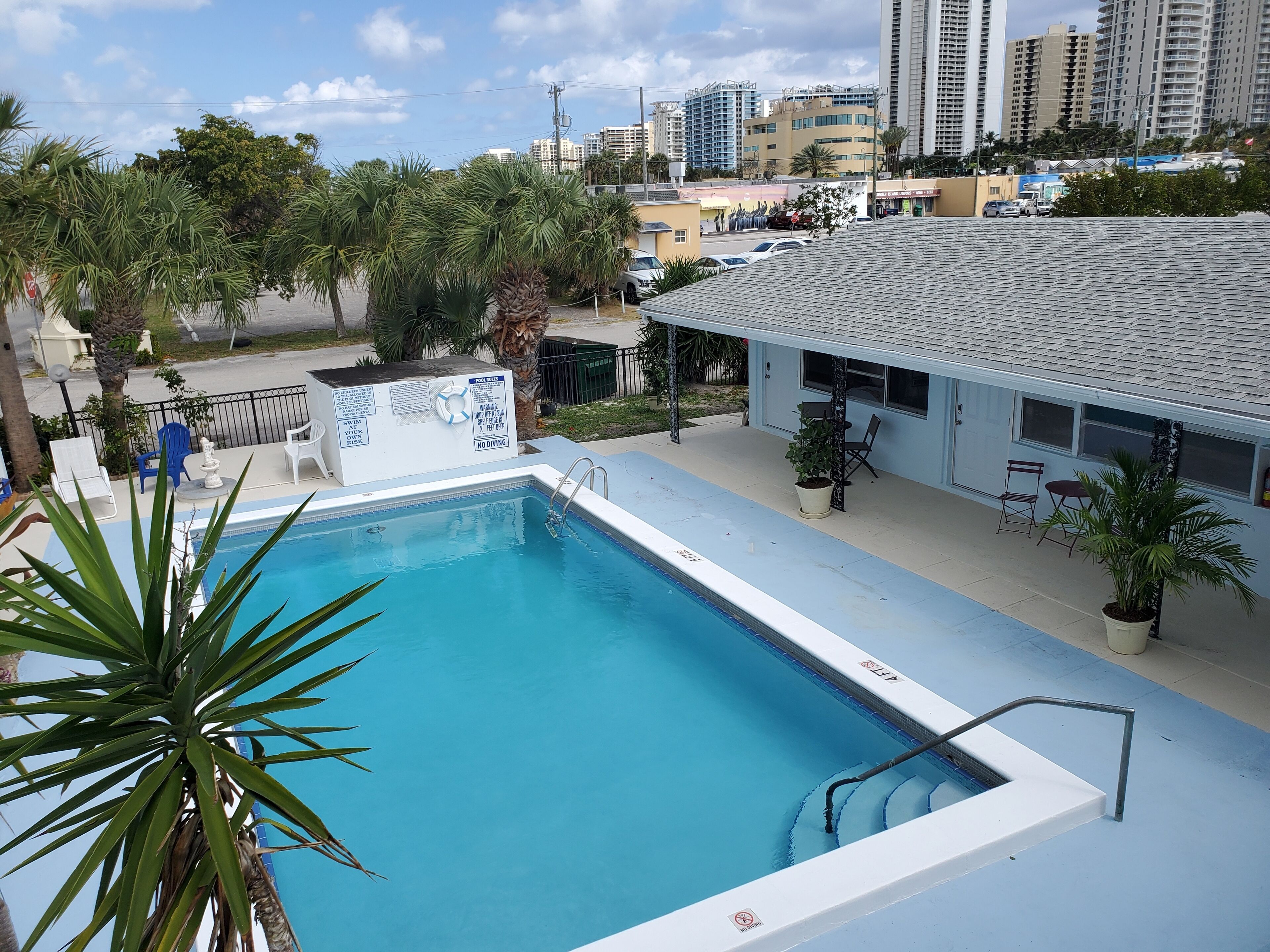 Una piscina con una escalera, rodeada de palmeras y un edificio con terraza.