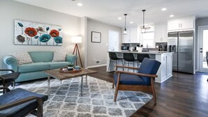 Ein modernes Wohnzimmer mit einer Couch, einem Couchtisch und einem Teppich. Im Hintergrund ist eine Küche mit Kühlschrank und Essbereich zu sehen.