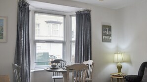 Ein Zimmer mit Esstisch und vier Stühlen, ein kleiner Beistelltisch mit Lampe, ein gerahmtes Bild an der Wand und ein Fenster mit Vorhängen.