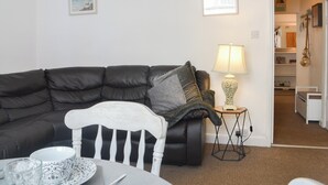 Ein Wohnzimmer mit einem dunkelbraunen Ledersofa, einem weißen Sessel, einem kleinen Tisch mit Schüssel und Löffel, einer Lampe und zwei gerahmten Bildern an der Wand.