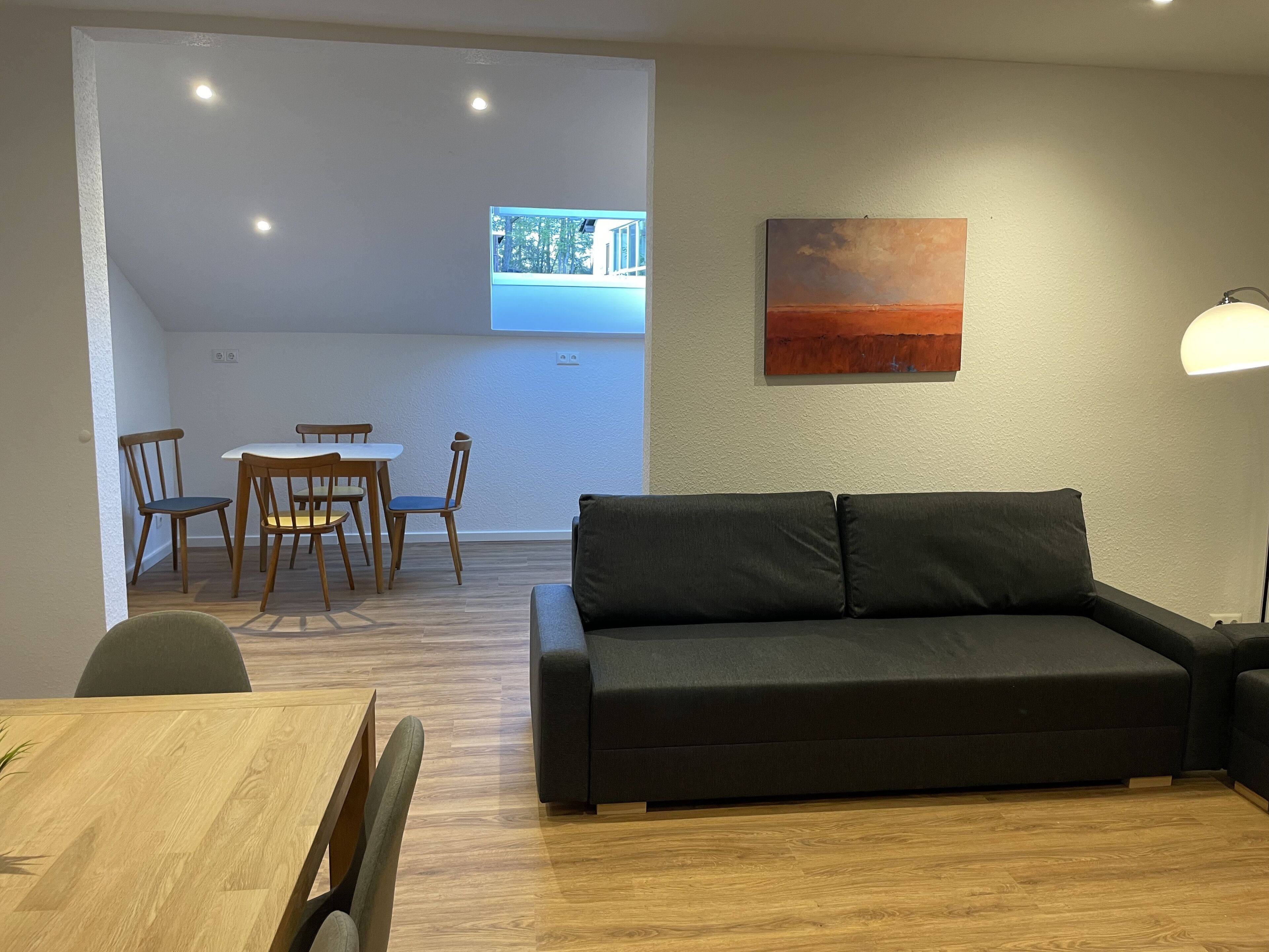 A modern living room with a dark sofa, wooden dining table, and chairs. There is a painting on the wall and a window with a view of trees.