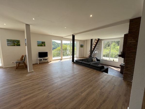 A spacious living area with a large sofa, a television on a stand, and a staircase leading to a balcony.