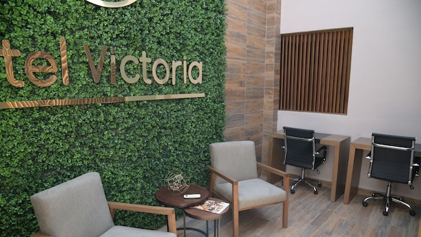 Zona de recepción de hotel con pared verde, mobiliario de madera y ventana con listones de madera.